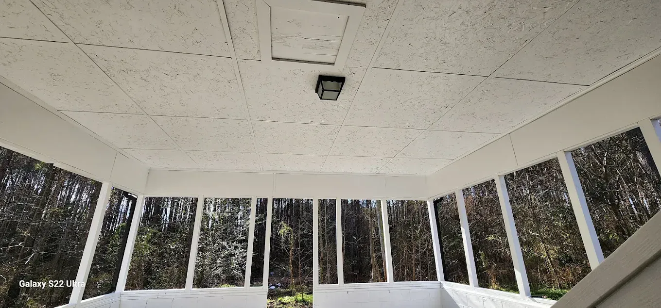 White sunroom ceiling with a light fixture and grid of panels, viewed from below looking out at a wooded area.