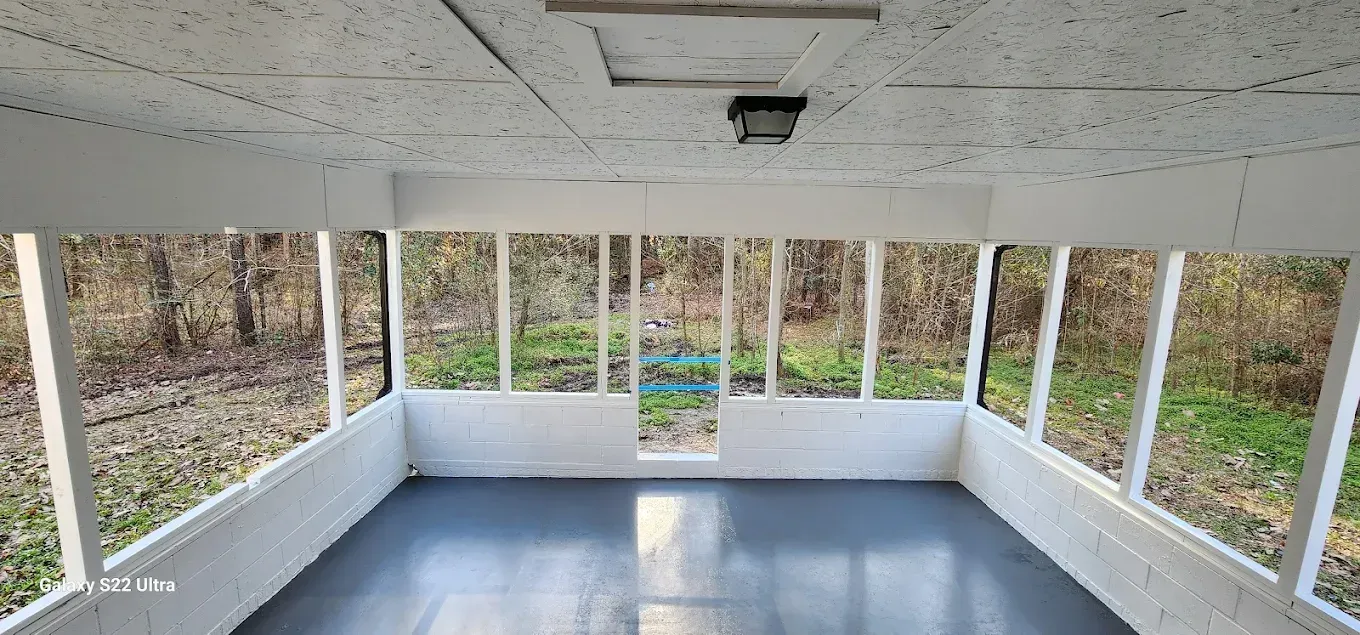 A brightly lit, enclosed porch with white walls, a gray floor, and large windows looking out onto a wooded area.