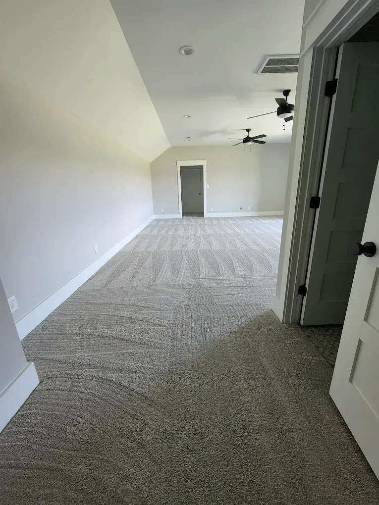 A spacious room with angled ceilings, light walls, tan carpet, two ceiling fans, and a door leading to another area.