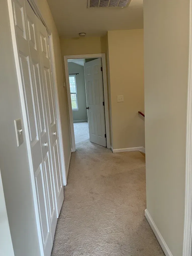 A view down a beige-carpeted hallway toward an open white door leading to another room with a window.