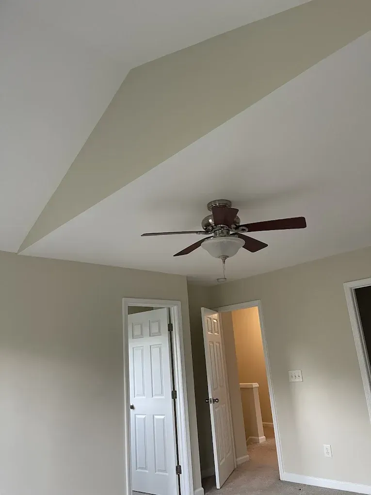 A room with beige walls, a slanted ceiling, and a ceiling fan, leading to a hallway with two white doors.