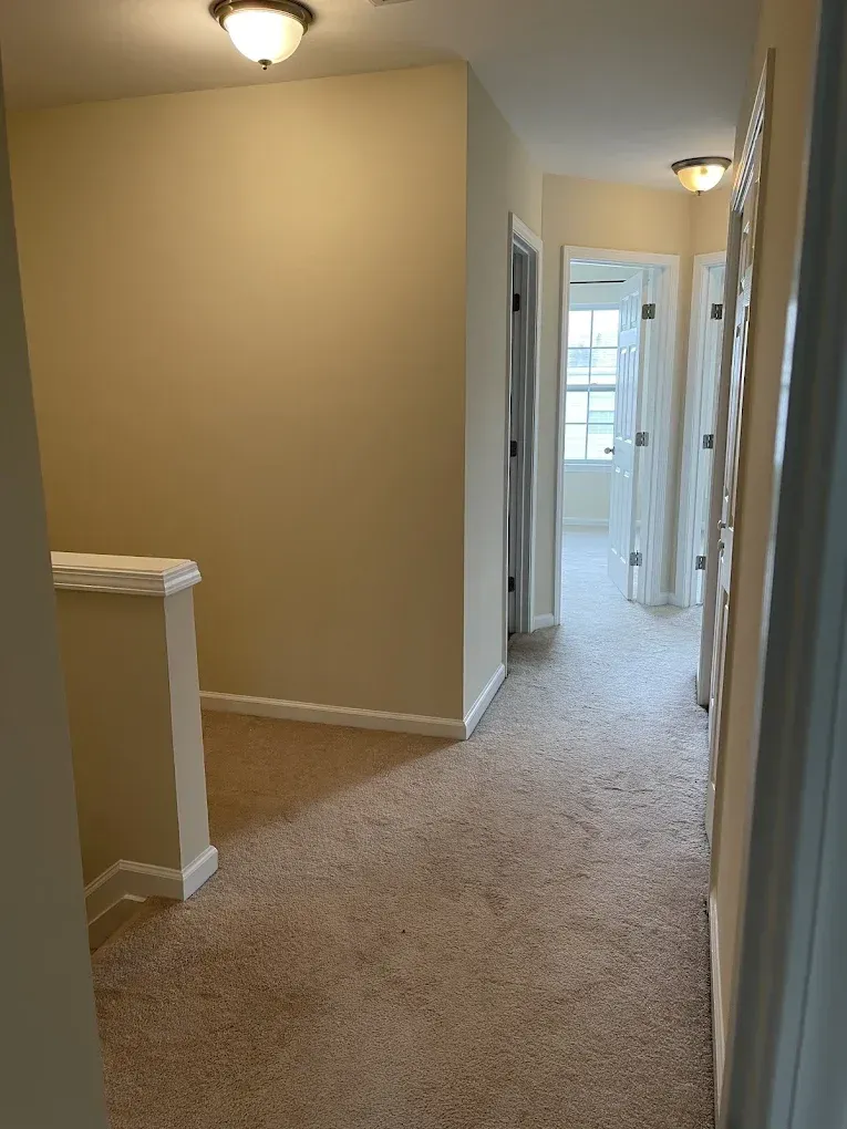 A carpeted hallway with beige walls, a small landing wall, and two open doorways leading to bright rooms.