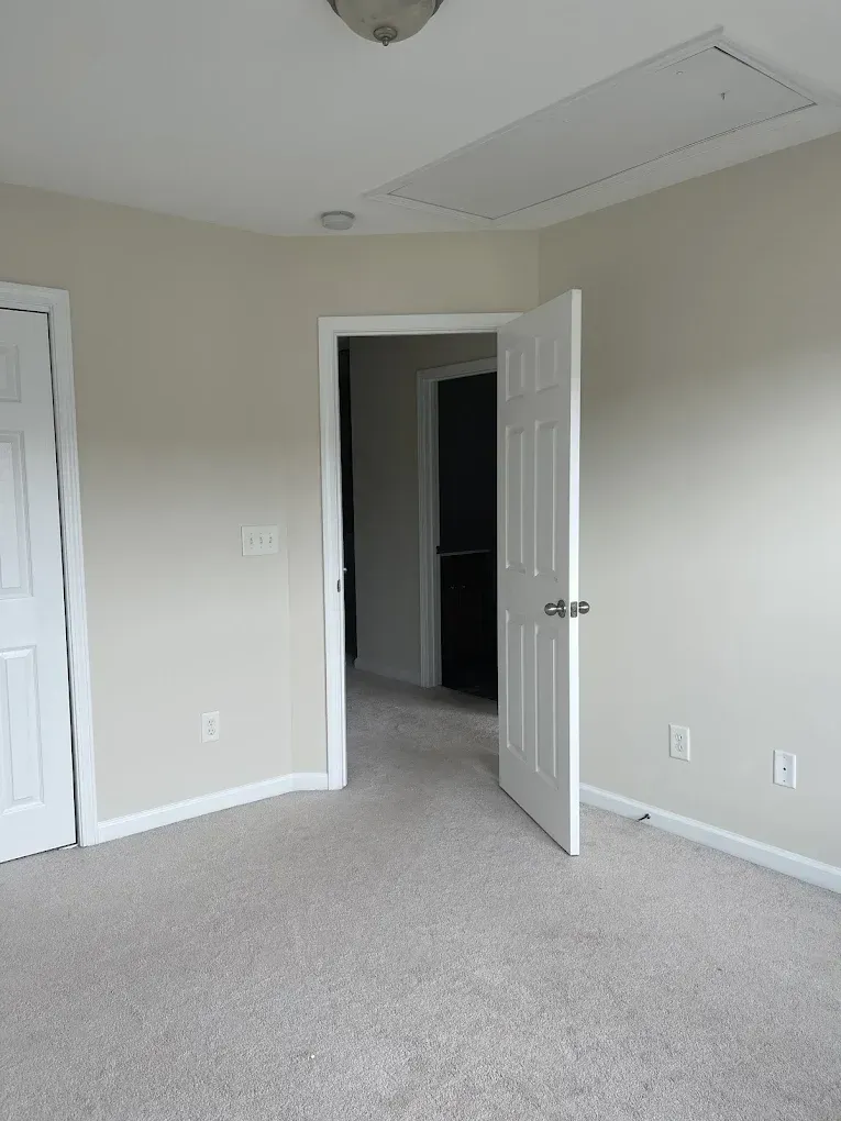An interior view of a room with light beige walls and carpet, showing two white interior doors and a ceiling attic hatch.