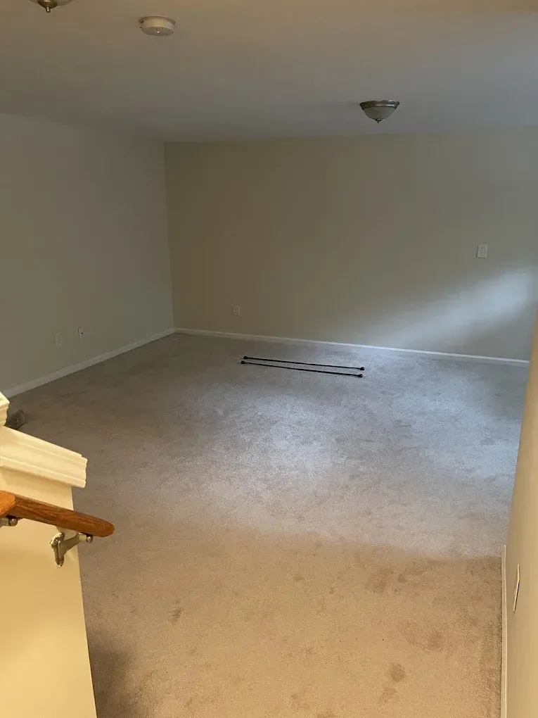 An empty, carpeted room with beige walls, a wooden stair railing in the foreground, and a floor vent in the center.