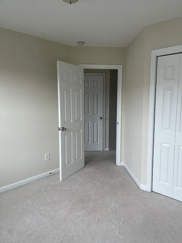 A neutral-toned room with beige walls and light carpeting, featuring an open door leading to a hallway.