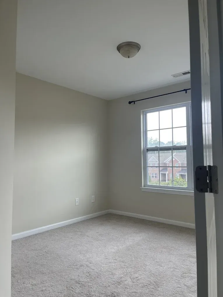 A view through a doorway into an empty bedroom with beige walls, gray carpeting, a window, and a ceiling light fixture.