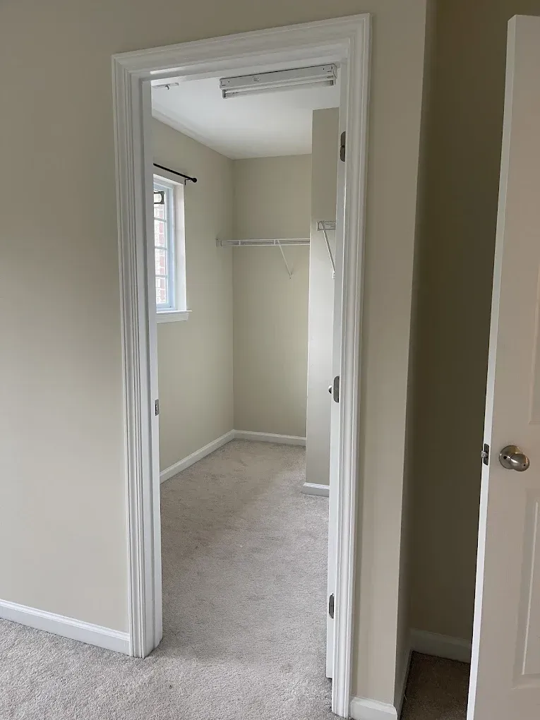 A view through a doorway into an empty walk-in closet with carpeted floors, beige walls, and a small window.