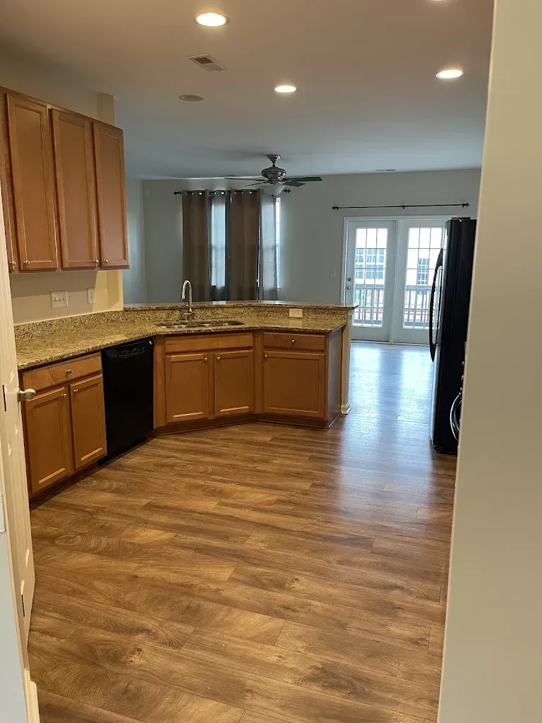 A kitchen with light wood cabinets, granite countertops, wood-look flooring, and a view into a bright living area.