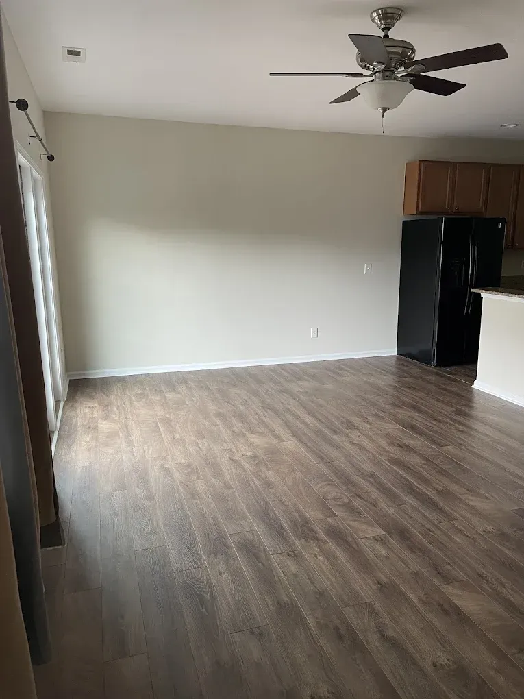 An open-plan room with wood-look flooring, a ceiling fan, a large empty wall, and a black refrigerator in the corner.