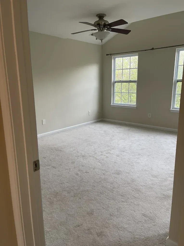 An empty bedroom with light-colored walls, cream carpeting, a ceiling fan, and two windows with a curtain rod.