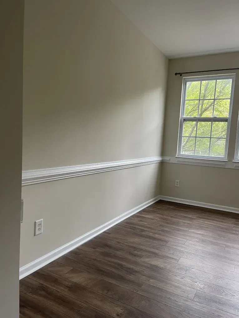 A view of a corner of a room with light beige walls, white chair rail and baseboard trim, and dark wood-look flooring.