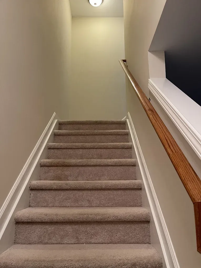 Staircase with light-colored carpet, beige walls, and a wooden handrail, viewed looking upward toward a light fixture.