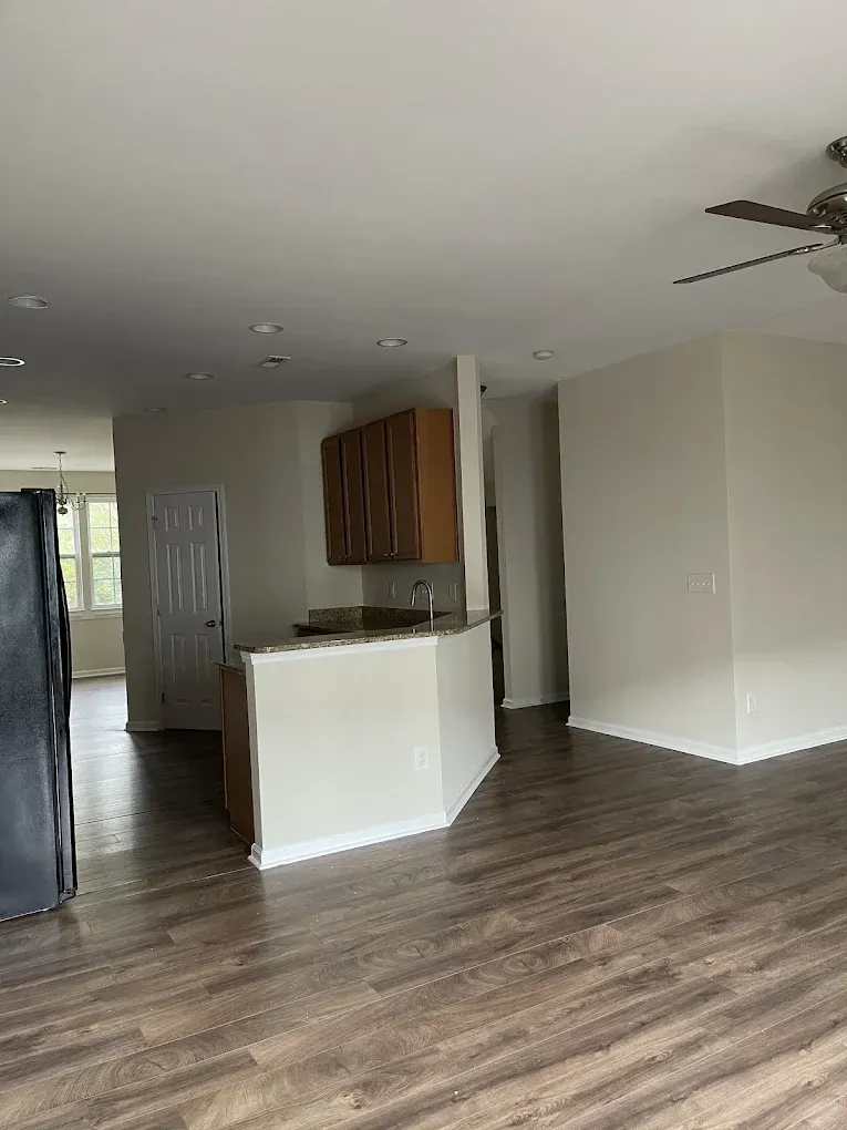 An open-plan interior showing a kitchen with wood cabinets, a peninsula counter, and light flooring leading to a doorway.