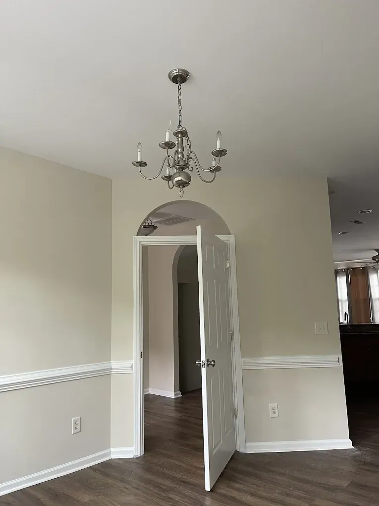 A white arched doorway with an open door leads to another room, under a chandelier in a room with beige walls and trim.