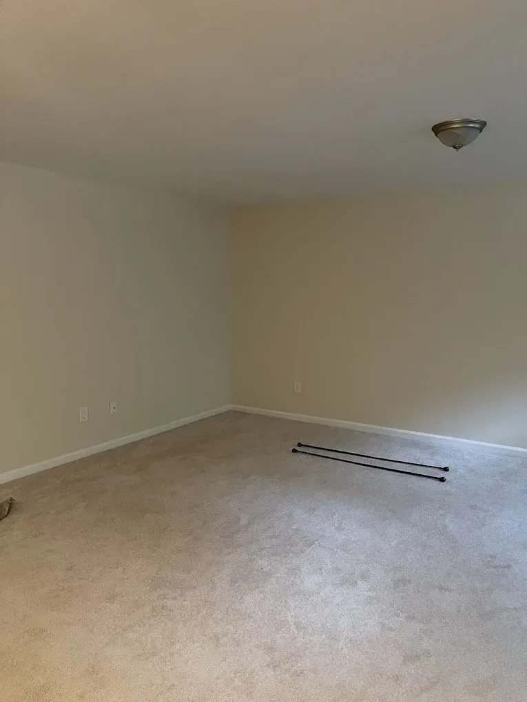 An empty room with beige carpet and walls, a ceiling light, and black markings on the floor.
