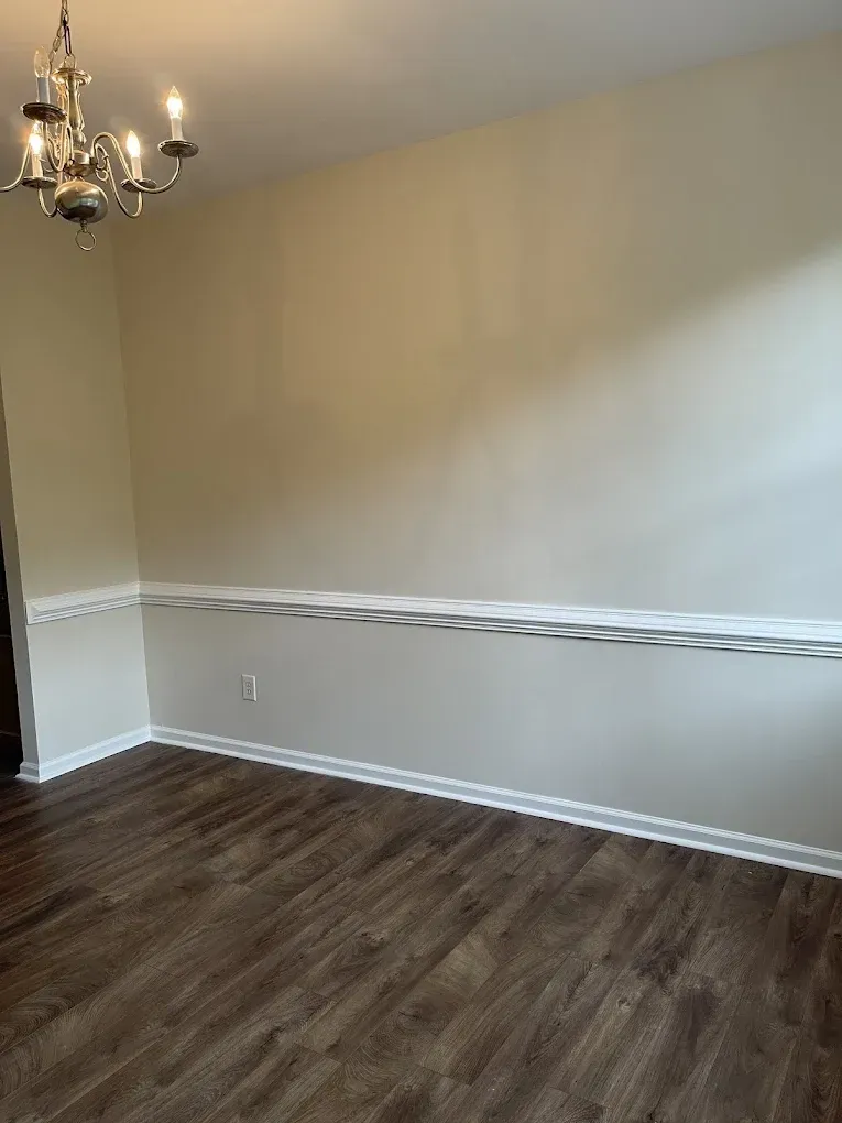 An empty room featuring dark wood-look flooring, light beige walls with white chair rail molding, and a chandelier.