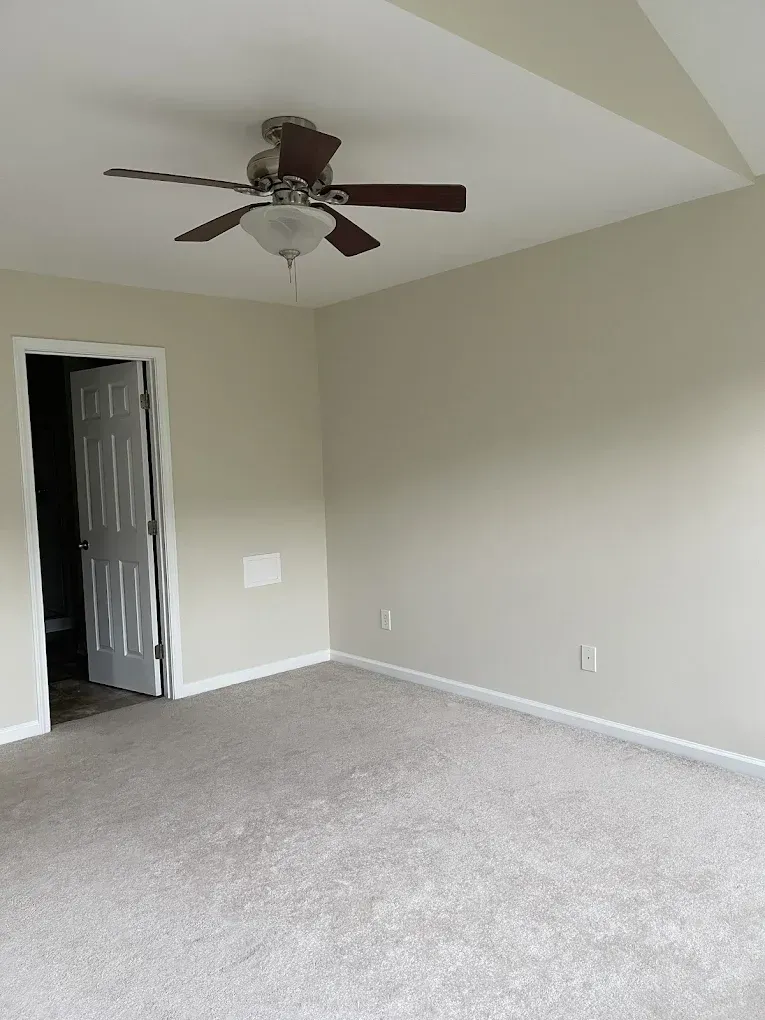 An empty room with beige walls, light carpet, a ceiling fan, and a white open door leading to an adjoining space.