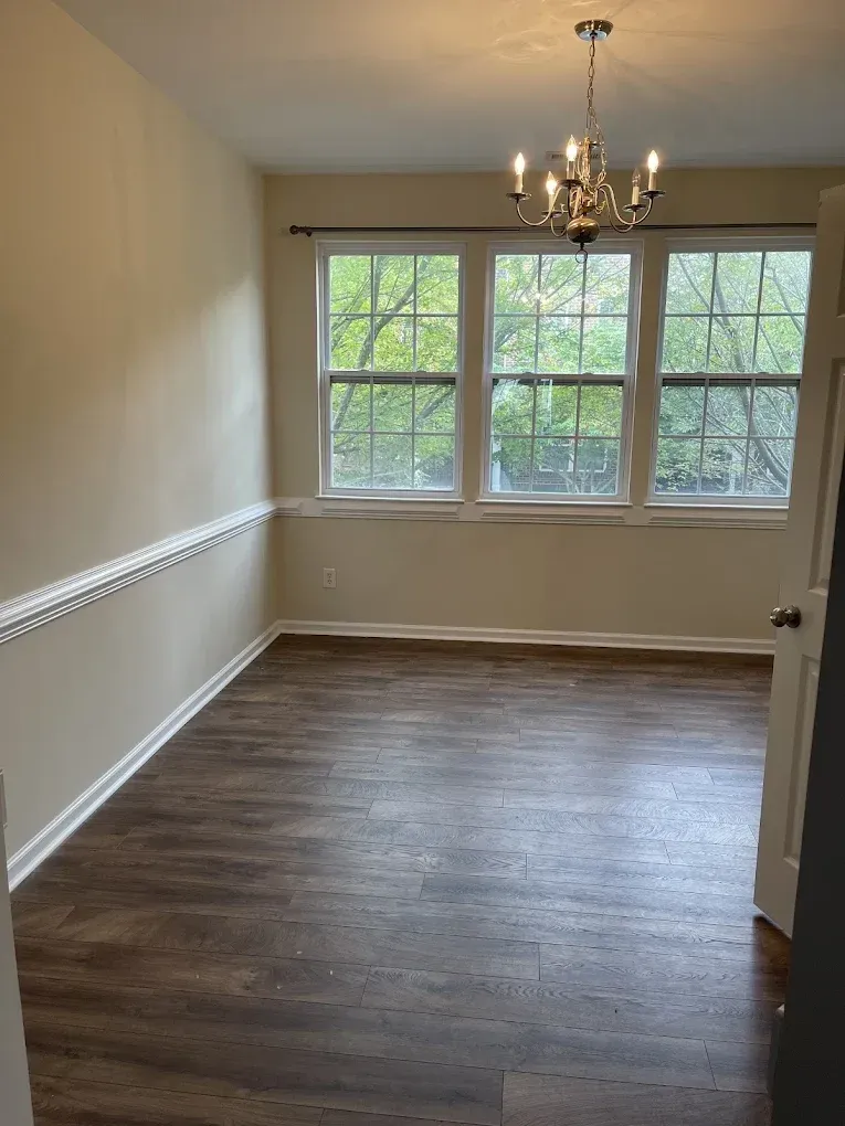 An empty dining room with tan walls, white chair rail molding, a chandelier, and dark wood-look flooring.