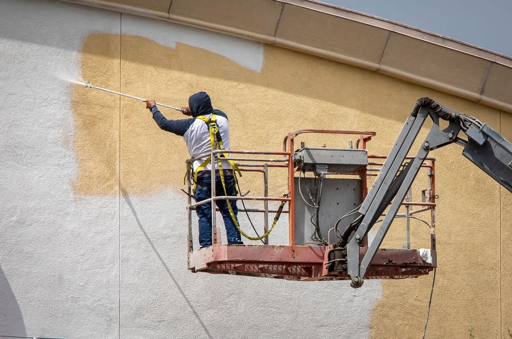 A worker in a safety harness uses a sprayer to paint a beige building wall from a raised aerial lift platform.