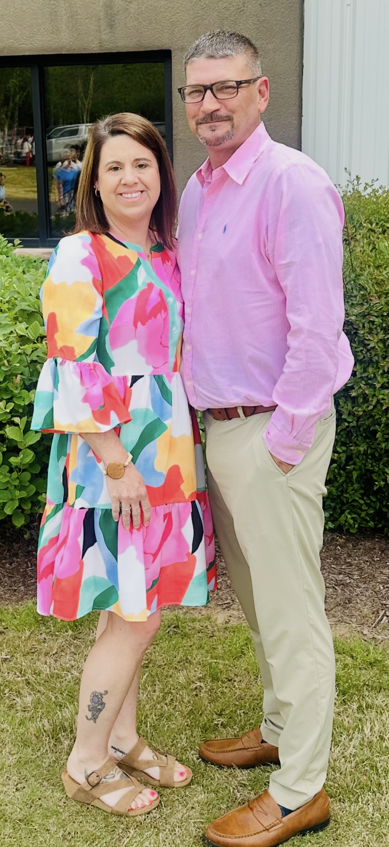 A man and a woman are standing next to each other in the grass . the woman is wearing a colorful dress.