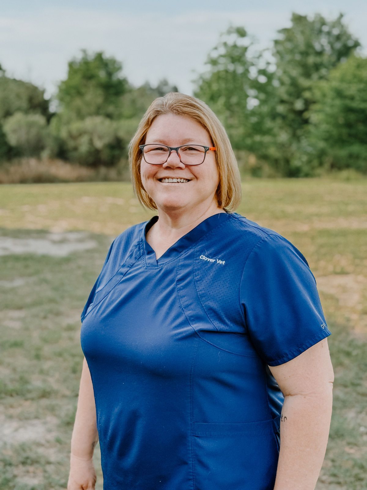 A woman wearing glasses and a blue scrub top is smiling in a field.