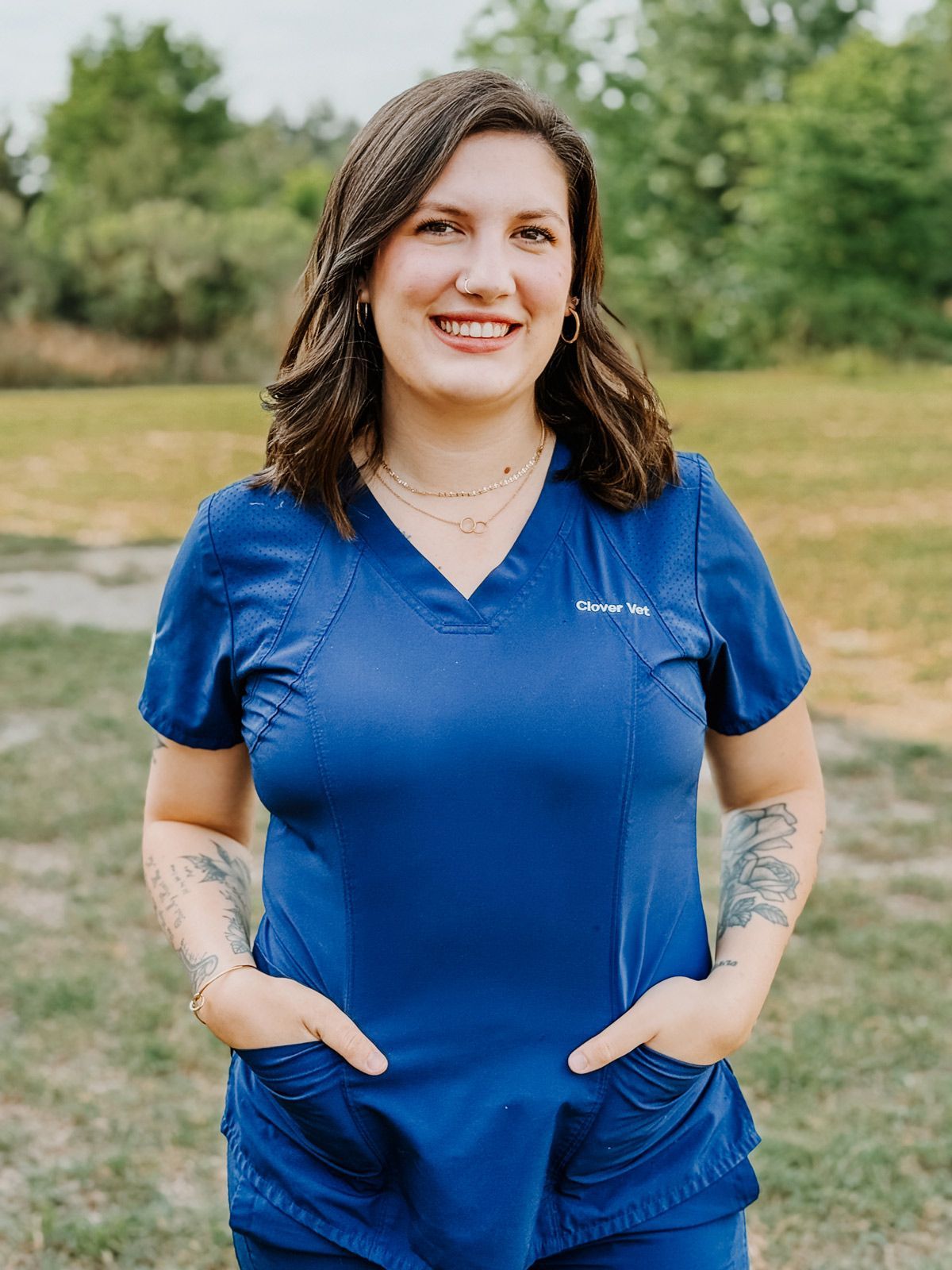 A woman in a blue scrub top is standing in a field with her hands in her pockets.