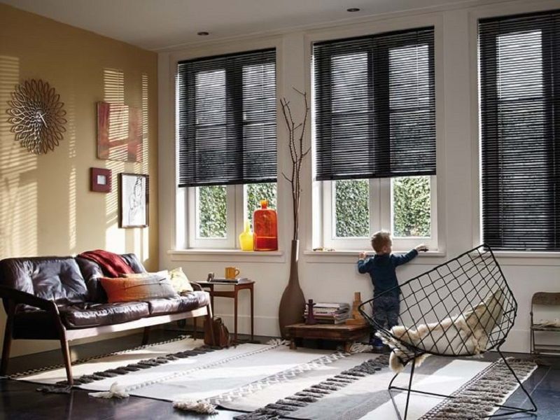 Living room with black blinds, child looking out window, and a dark brown sofa. Living room with black blinds, child looking out window, and a dark brown sofa.