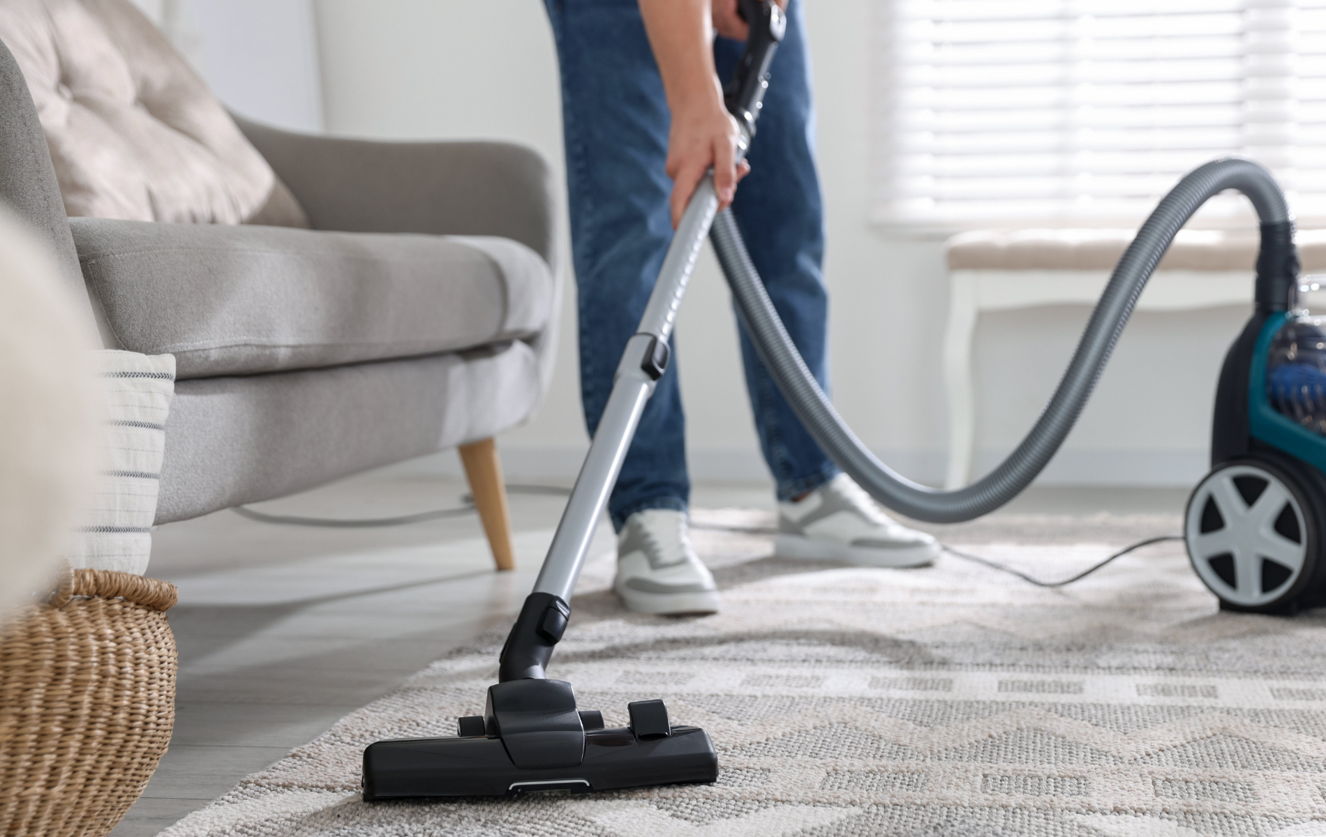 A person vacuums a patterned area rug in a brightly lit living room near a gray sofa.