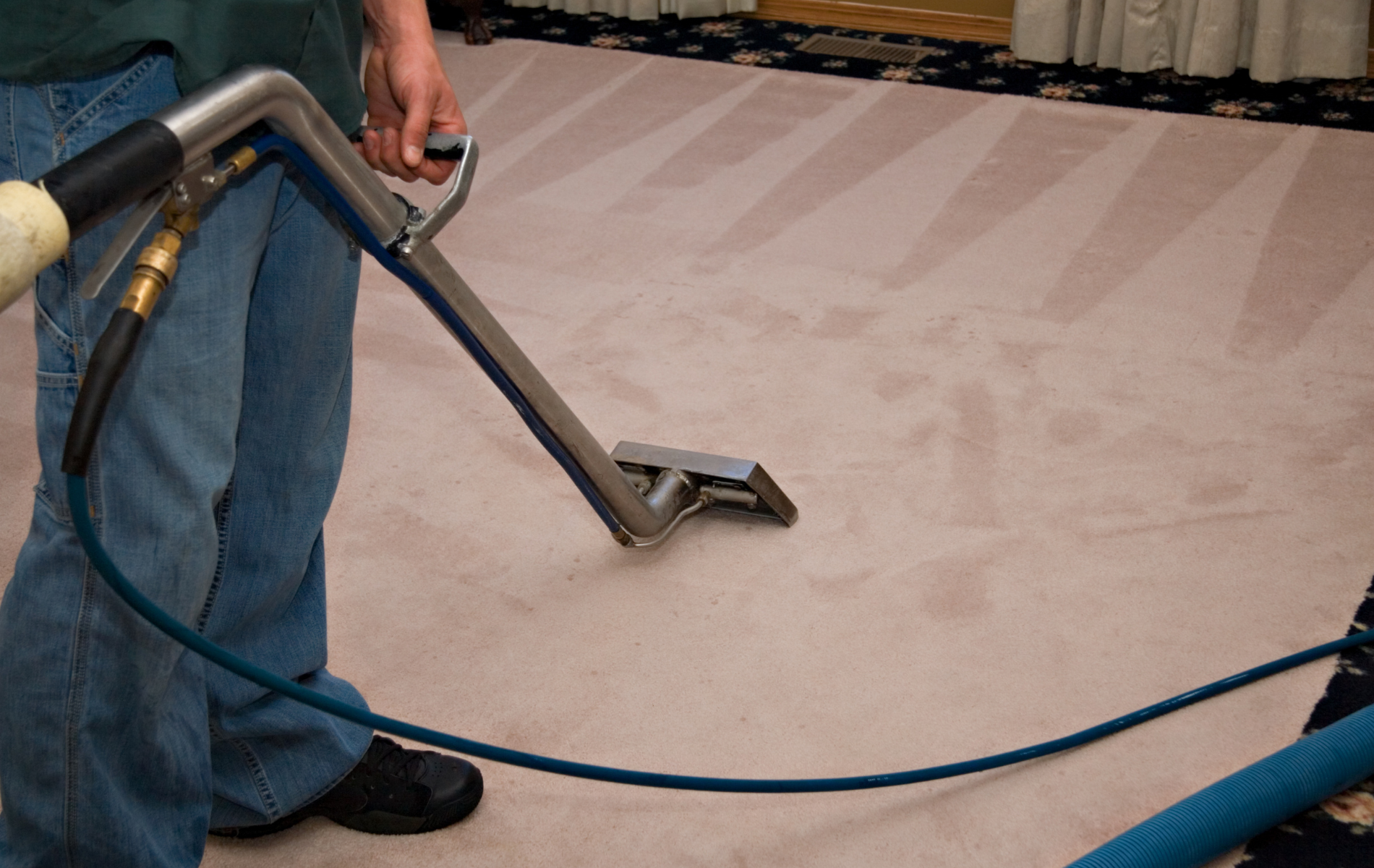 A person in jeans uses a metal carpet cleaning wand on light-colored carpet with visible cleaning tracks.