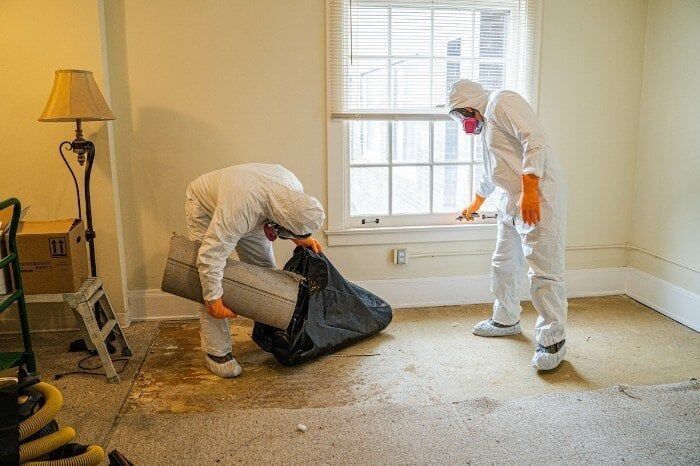 Two people in white protective suits and masks remove carpeting from a room during a cleanup or renovation project.