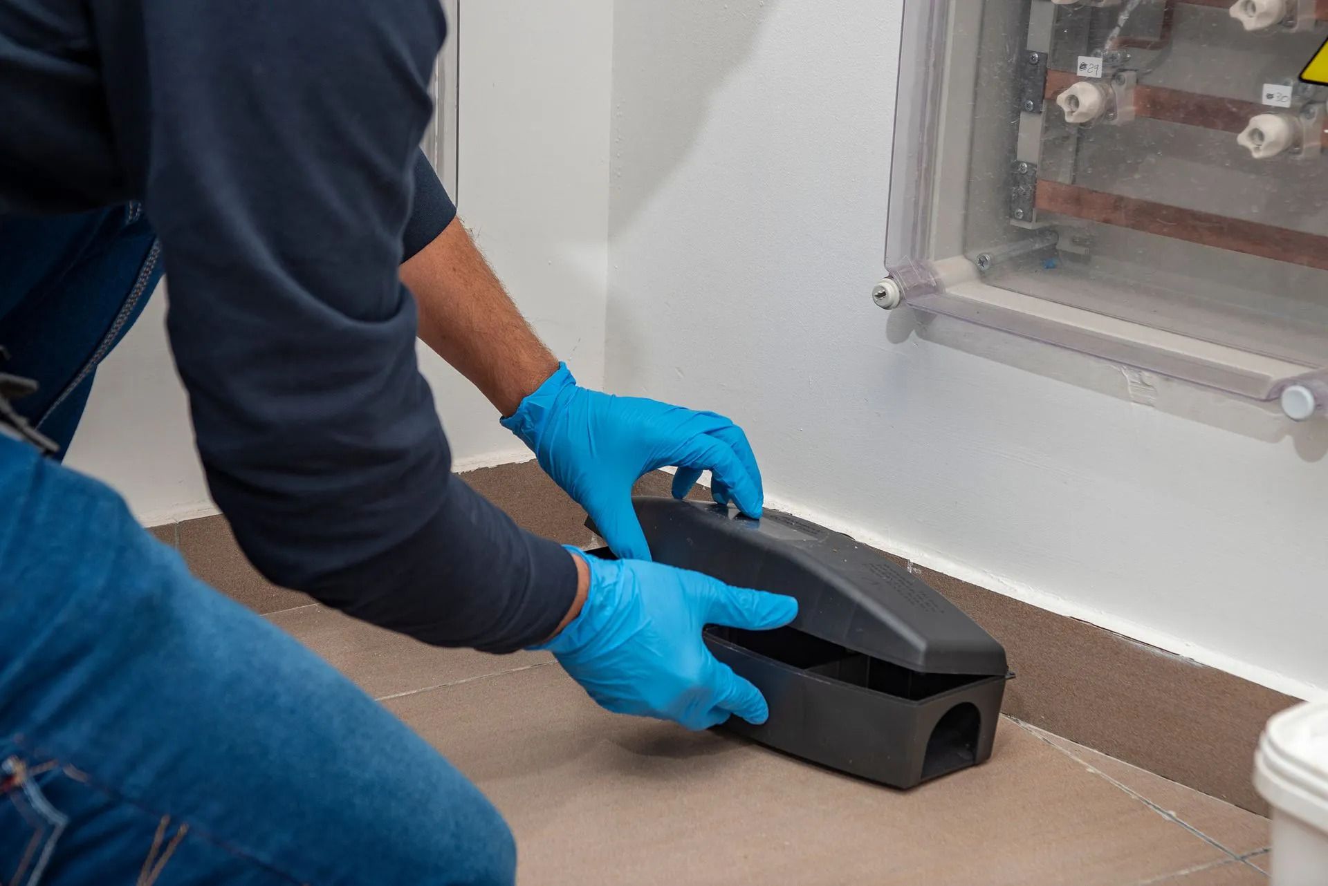 Person in blue gloves placing a black device on the floor near a wall-mounted electrical panel.