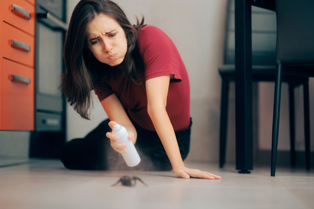 A woman crouched on the floor in a kitchen, looking concerned as she sprays insecticide at a large c