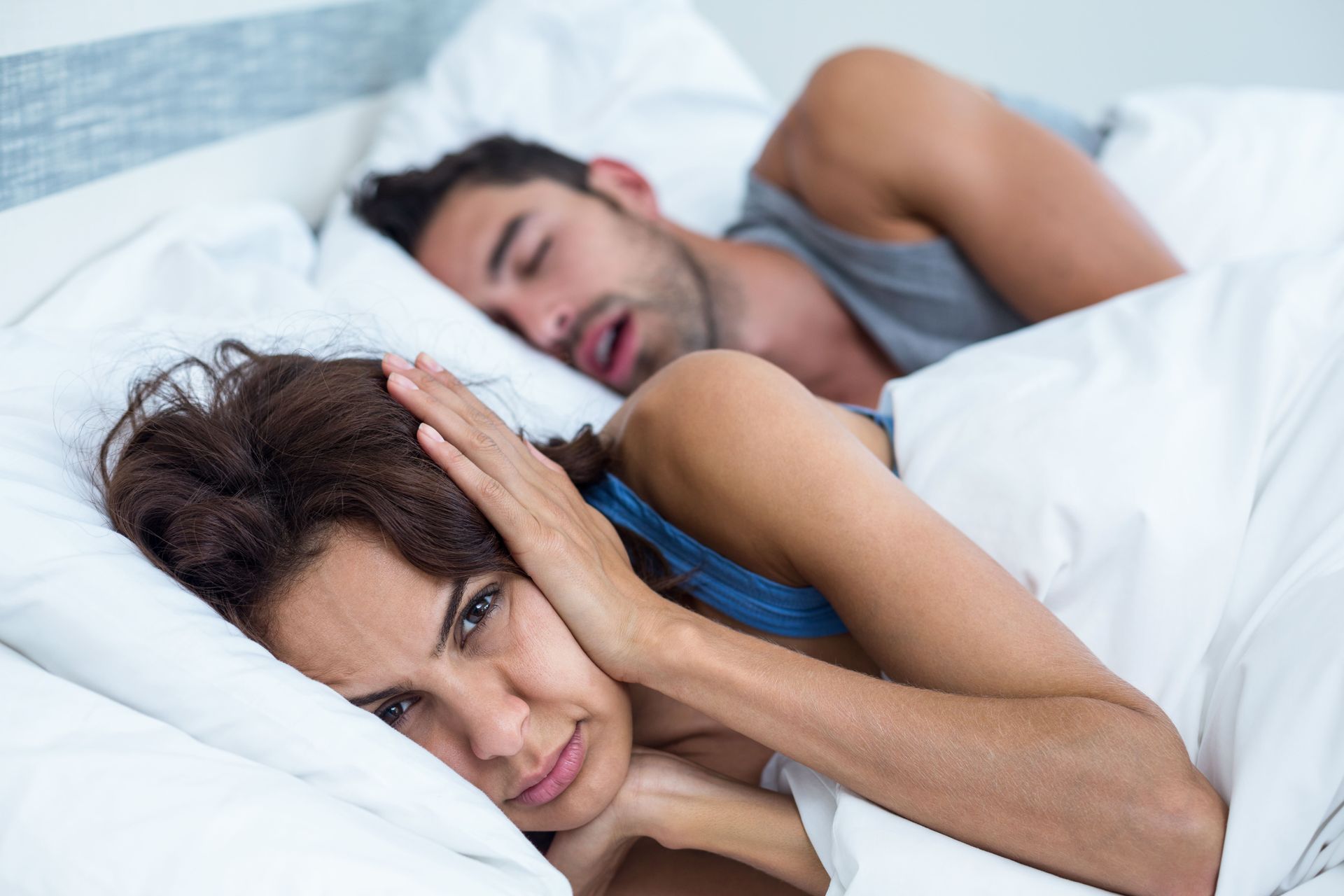 Duas pessoas deitadas na cama com travesseiros: uma está acordada e preocupada, enquanto a outra dorme atrás dela.