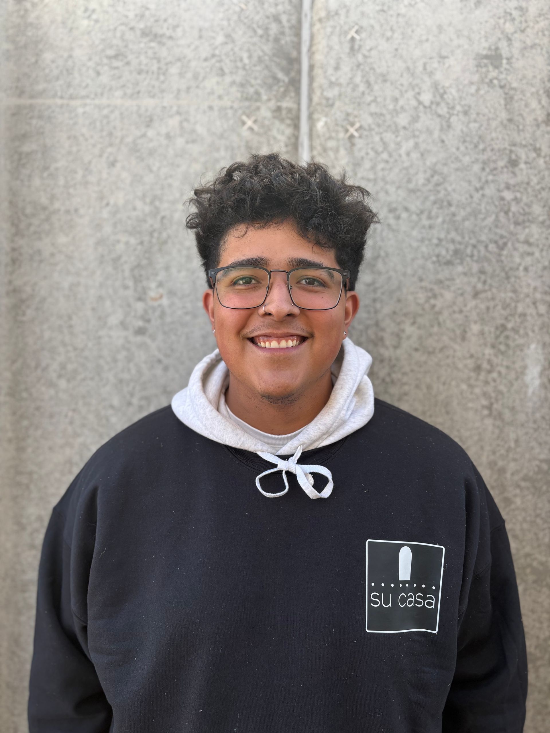 Person wearing glasses and a black hoodie smiles in front of a concrete wall.