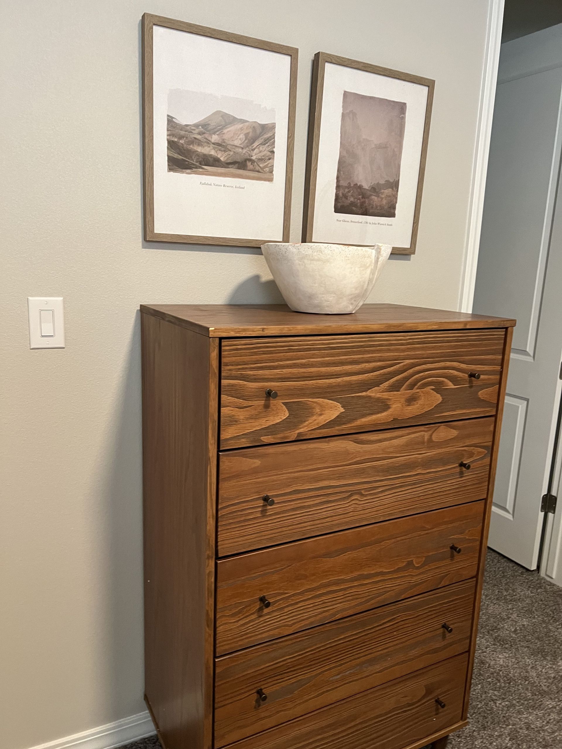 A wooden chest of drawers with two pictures on the wall above it.