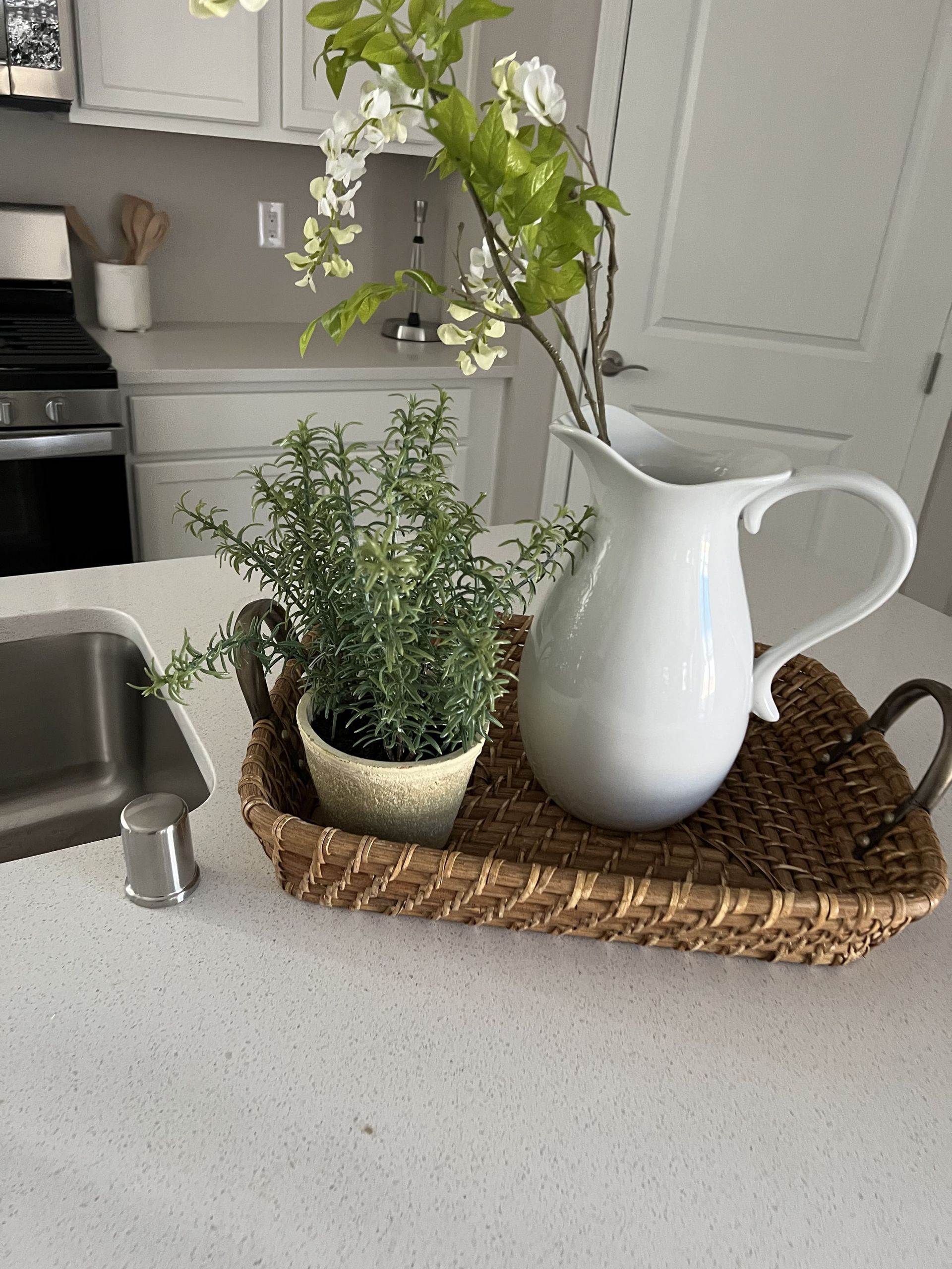 A pitcher and a potted plant are on a wicker tray on a kitchen counter.