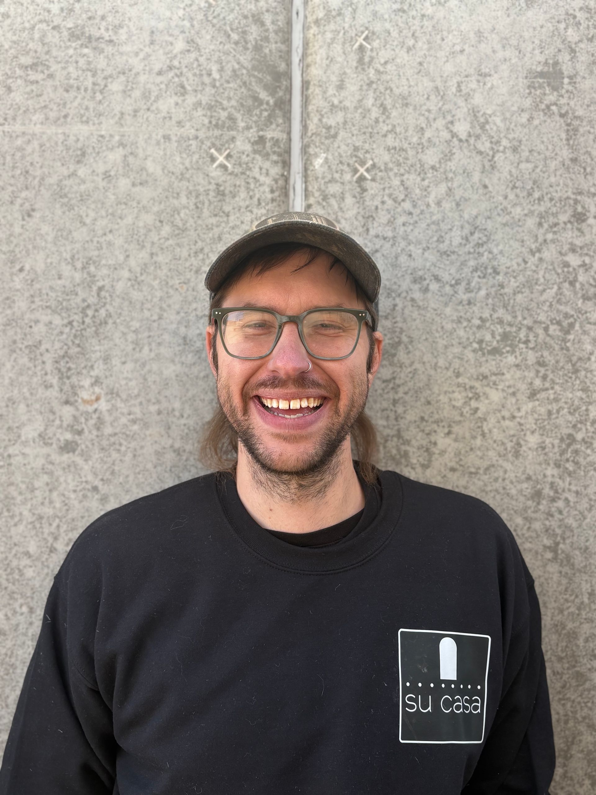 Man with glasses and cap smiles in front of a gray wall, wearing a black sweatshirt with a logo.