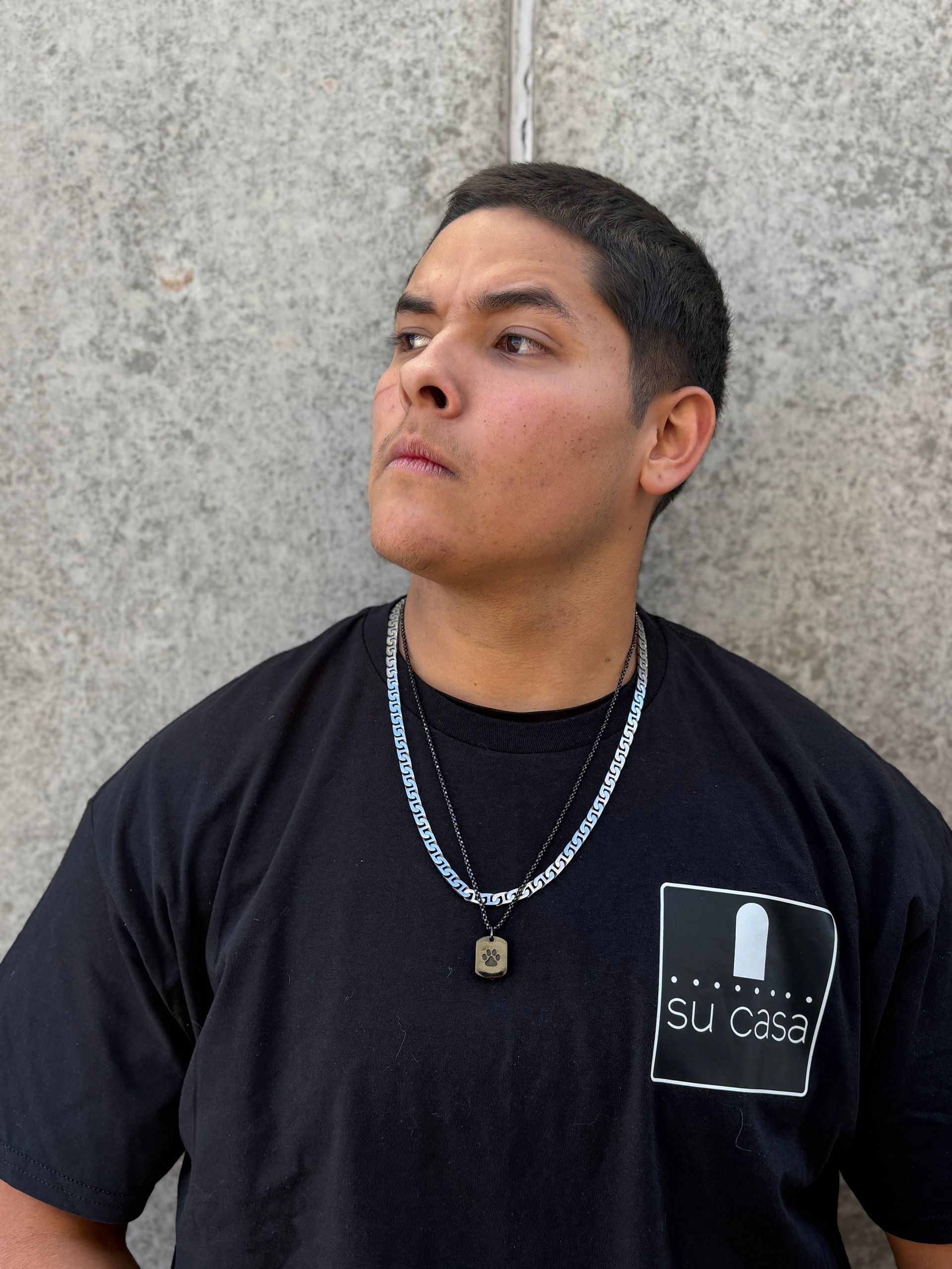 Man wearing a black t-shirt and two necklaces, looking upward, standing against a gray wall.