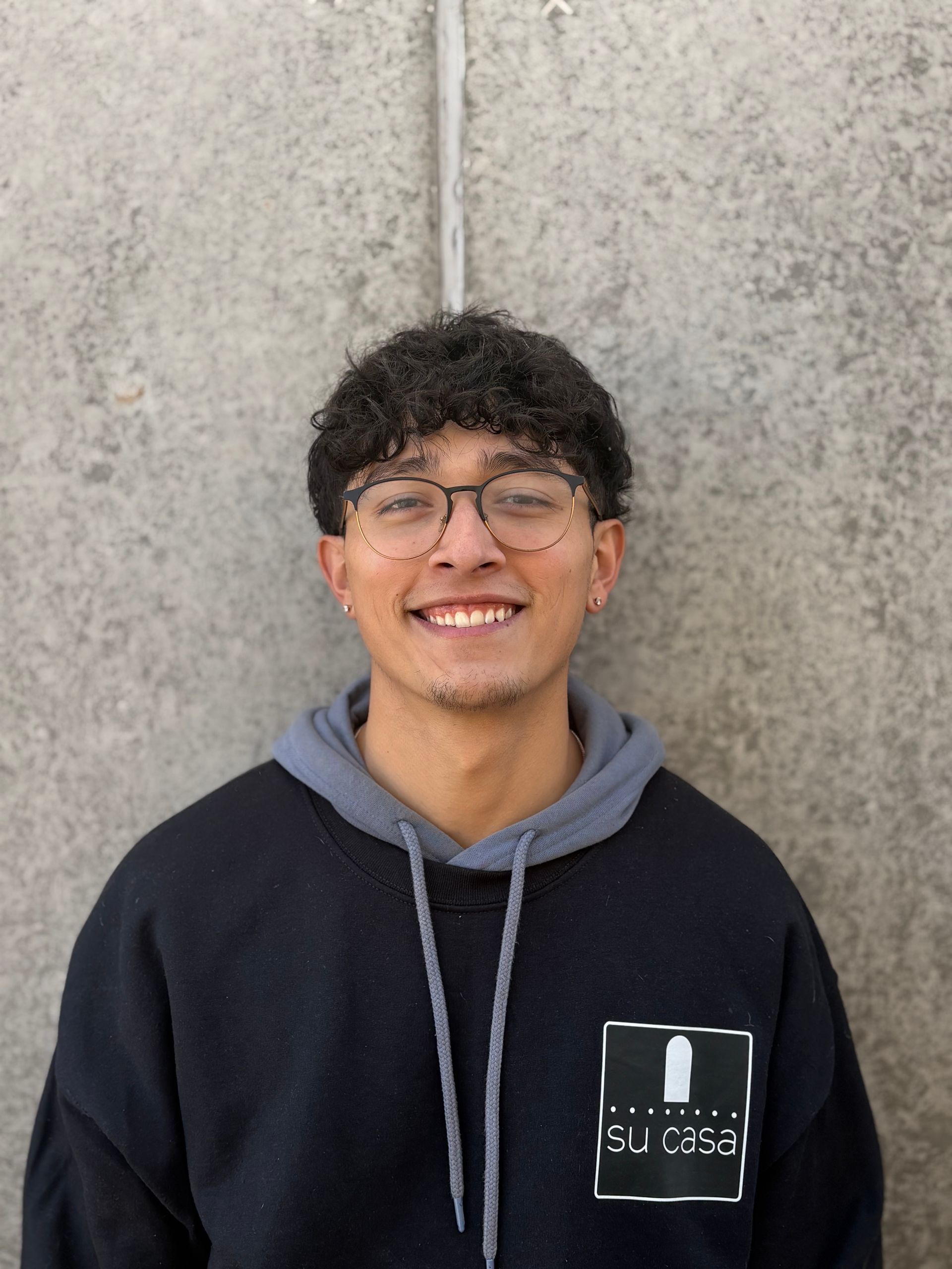 Person smiling, wearing glasses and a black hoodie with gray trim, standing in front of a concrete wall.