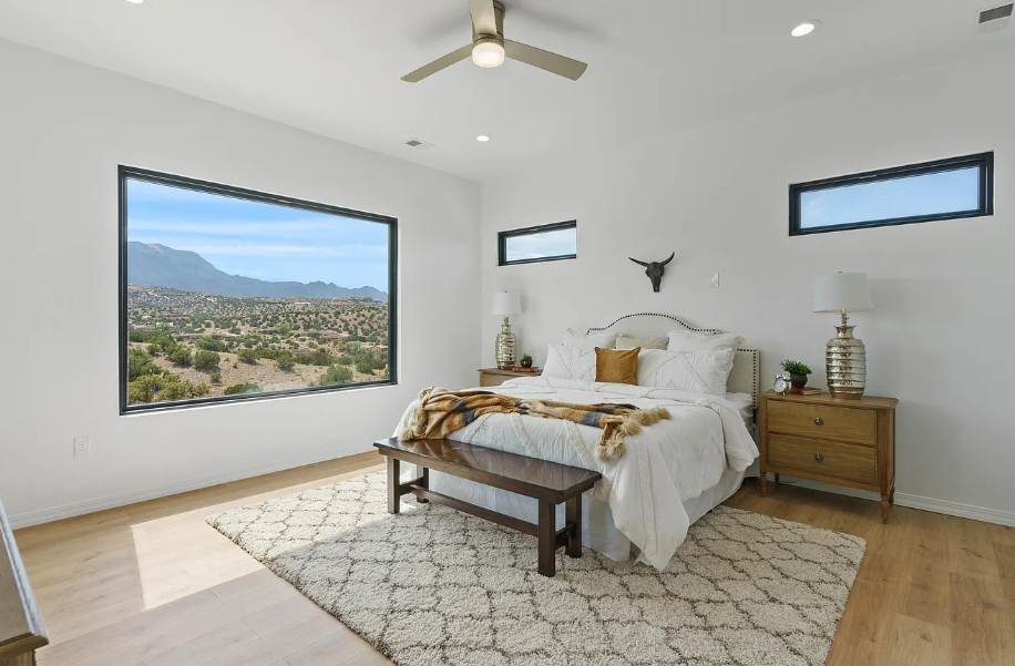A bedroom with a bed , a bench , a ceiling fan and a large window.