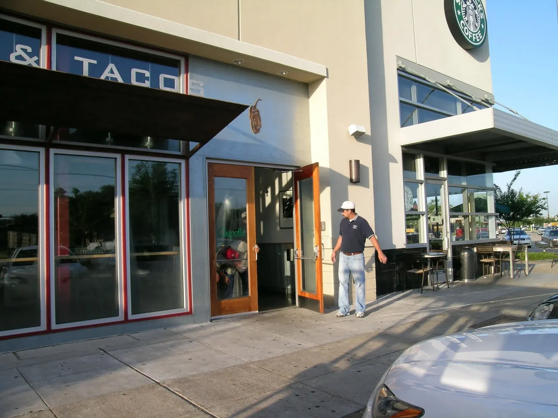 Taco restaurant entrance with open doors and a man standing outside by the sidewalk.