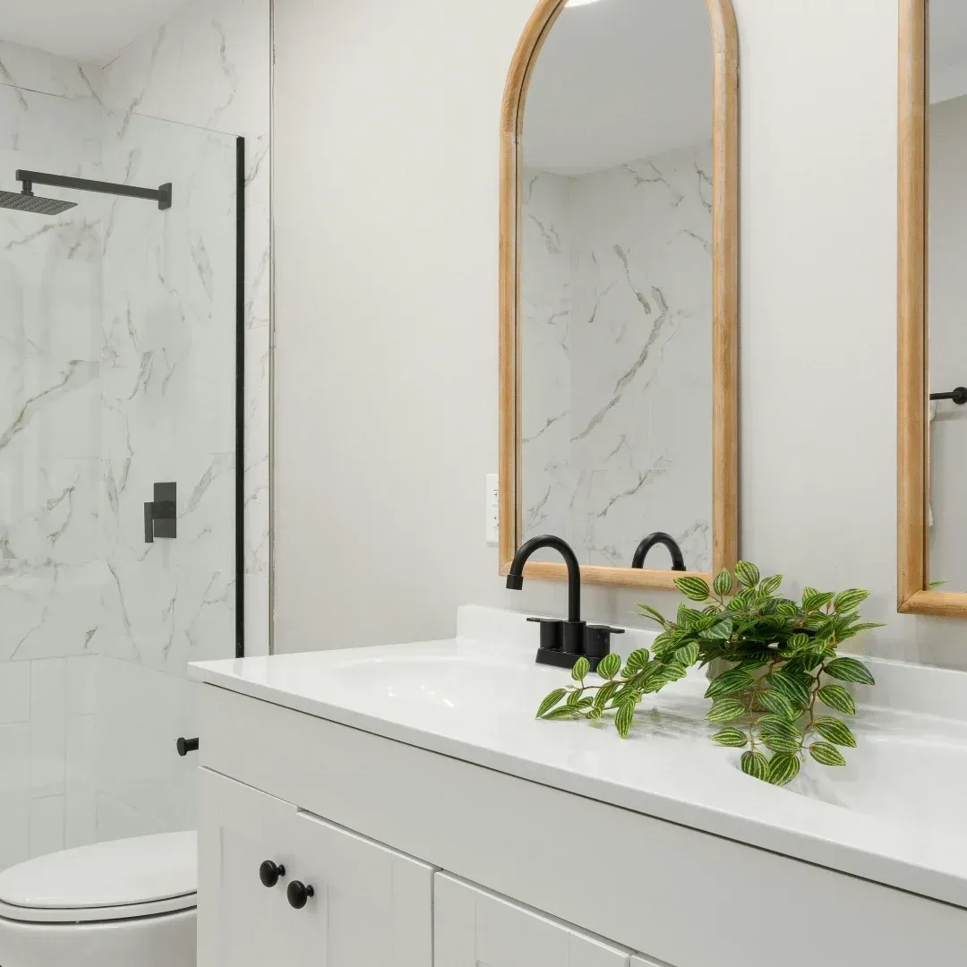 Modern bathroom vanity with arched mirror, black fixtures, white marble surfaces, and a small green plant