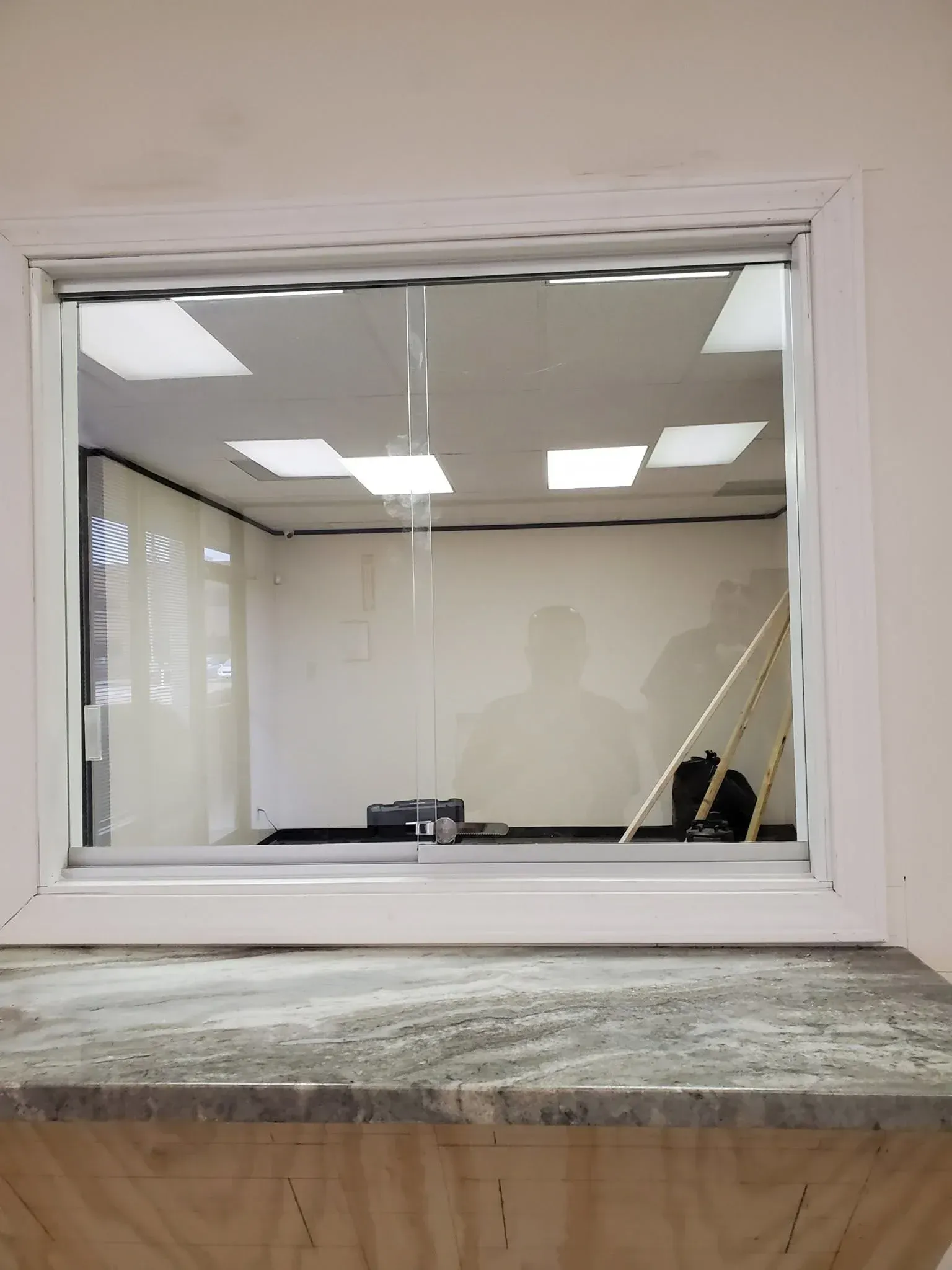 White-framed office window with blinds, showing an empty room and ceiling lights.