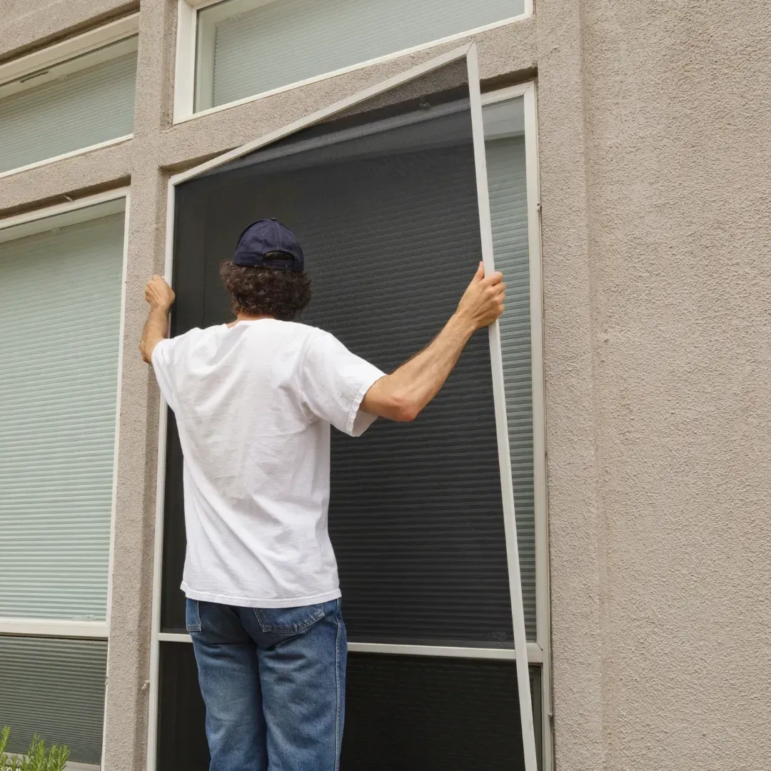 Person installing a white window screen in a beige exterior wall.