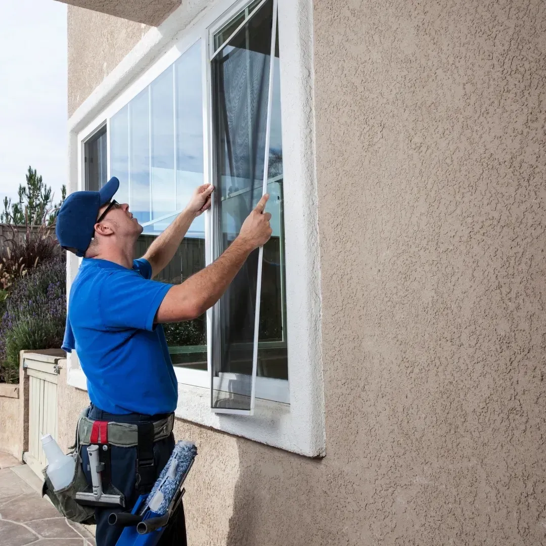 Man installing a window screen on a house exterior, wearing a blue shirt and cap