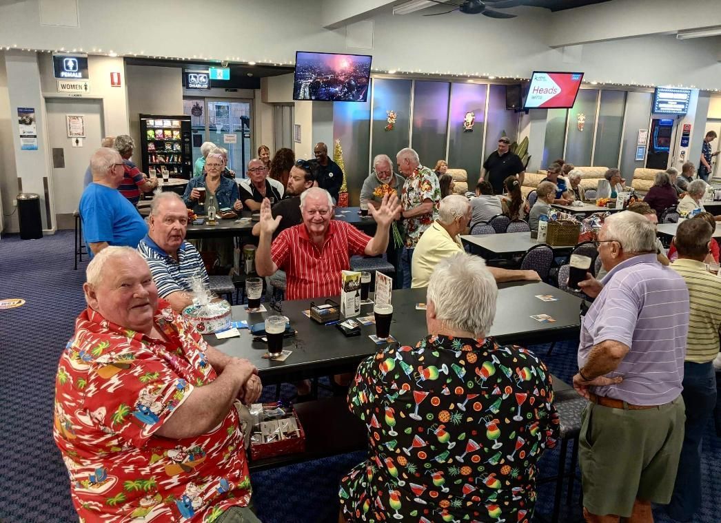 People gathered at tables in a club, some waving and smiling, drinks on tables, TVs on walls.