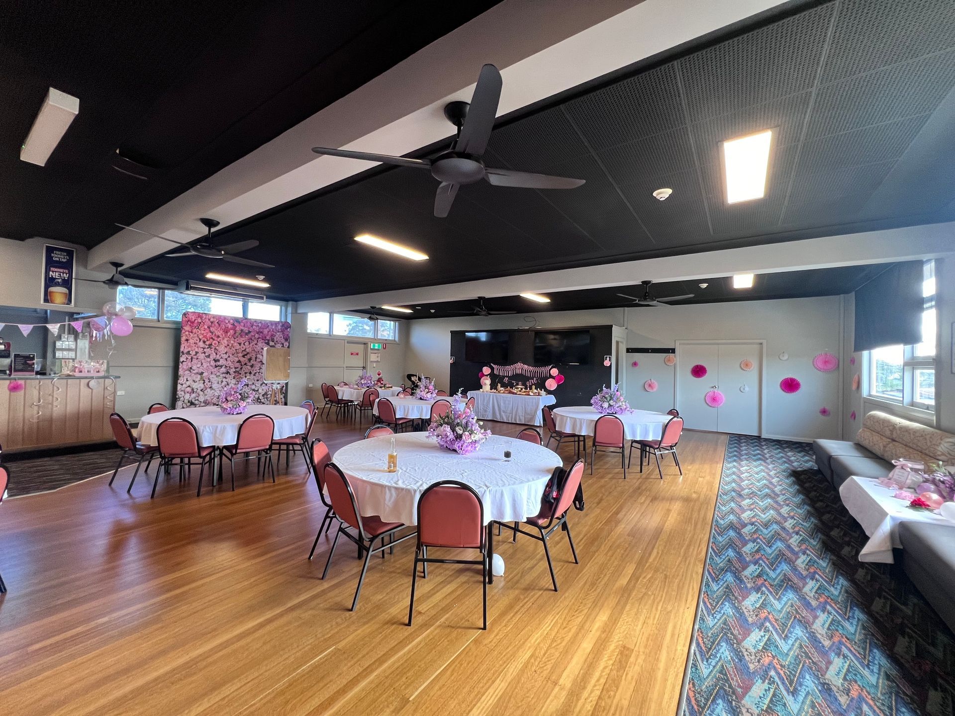 Empty banquet hall with round tables, pink chairs, wood floors, and ceiling fans under bright lights — Taree Railway Bowling Club Ltd In Taree, NSW