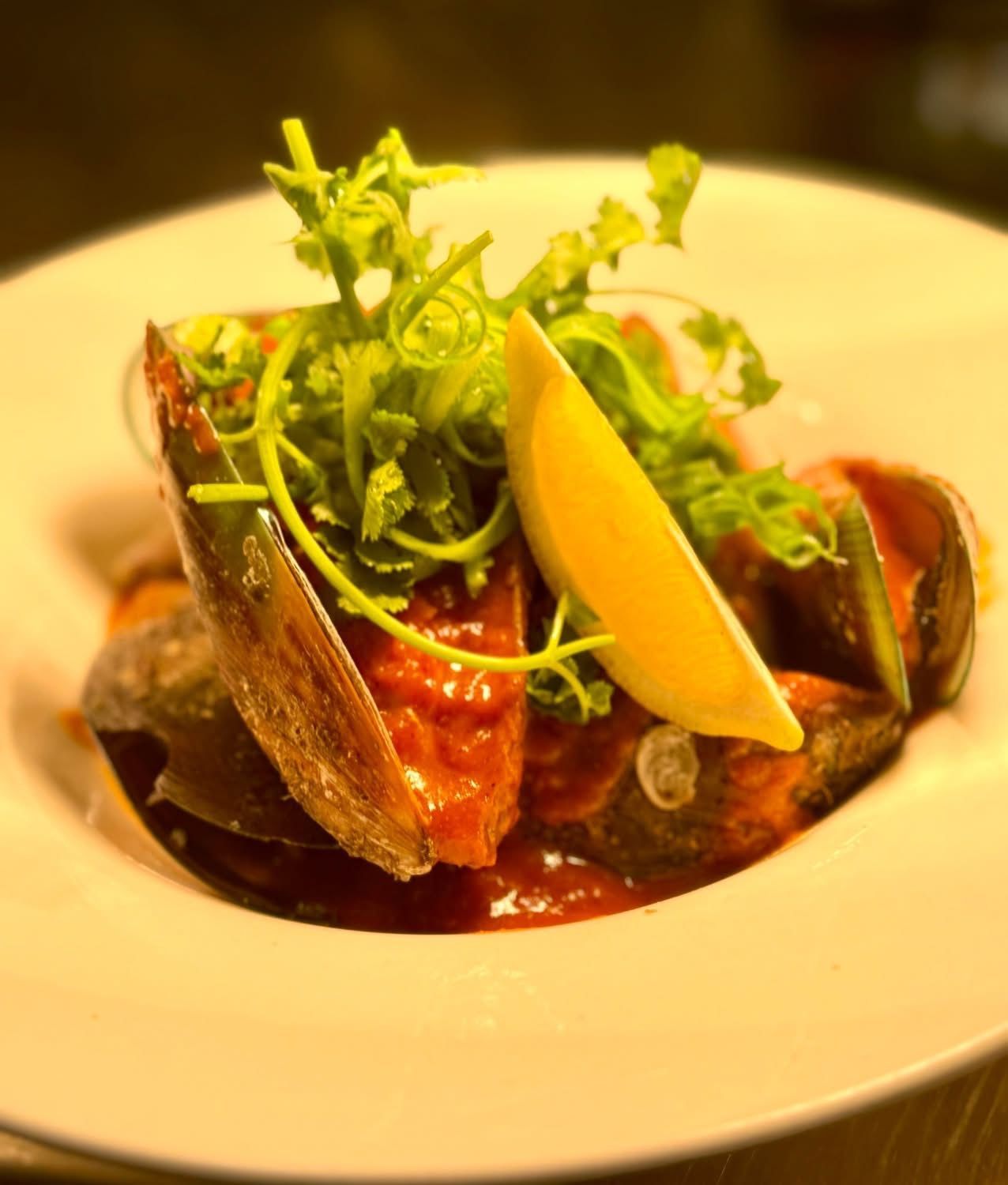 Mussels in tomato sauce, garnished with greens and lemon wedge, served in white bowl.