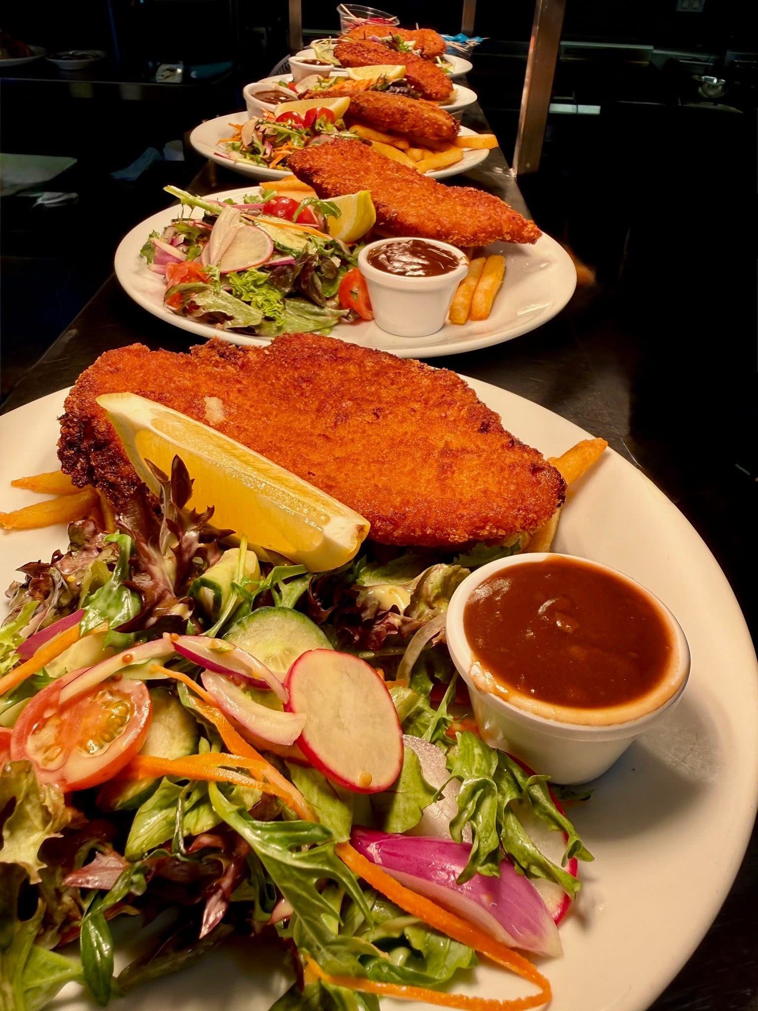 Plated schnitzel meals with salad, fries, lemon, and dipping sauce, arranged in a row on a black surface.