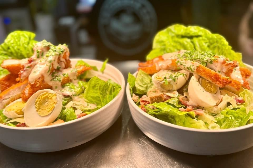 Two Caesar salads with fried chicken, eggs, and bacon in white bowls.